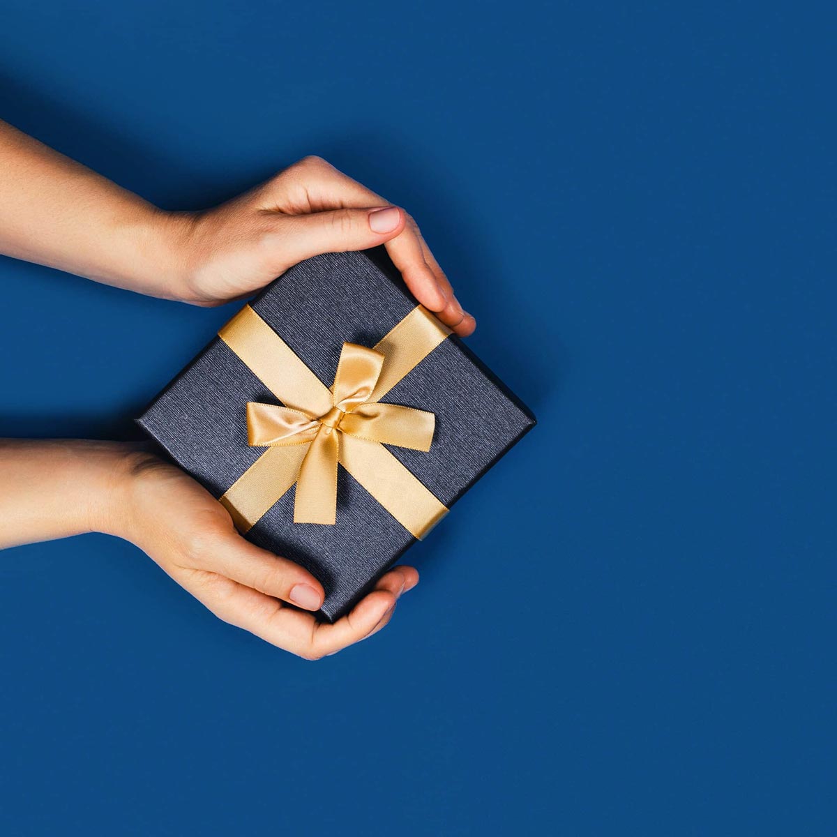 Une personne tient un paquet cadeau jaune or entre ses mains devant un fond bleu 